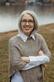 A white woman with platinum, chin-length hair and glasses is smiling confidently at the camera with her arms crossed and tucked in front of her.  She is wearing a white blouse with a tan parka sweater over it.  She is standing outside on a grassy hill with a partial view of a lake behind her.  